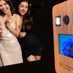 Photobooth Les Instantanes de Fred en bois clair installe a Arlon, avec trois jeunes femmes souriantes posant avec des accessoires fun