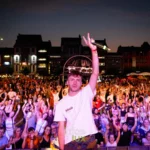 ALAN.V, micro en main et doigts en V, salue la foule lors du Bastogne Summer Festival 2025, de nuit, sur la place de Bastogne