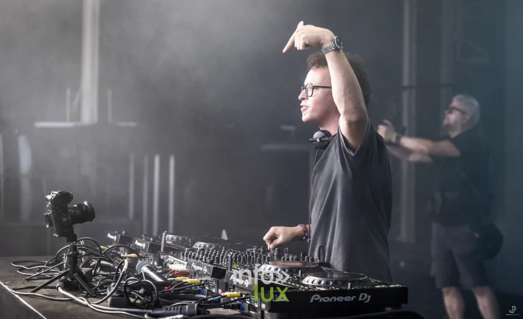 Un DJ à lunettes lève le bras au-dessus de sa table de mixage, dans une lumière blanche