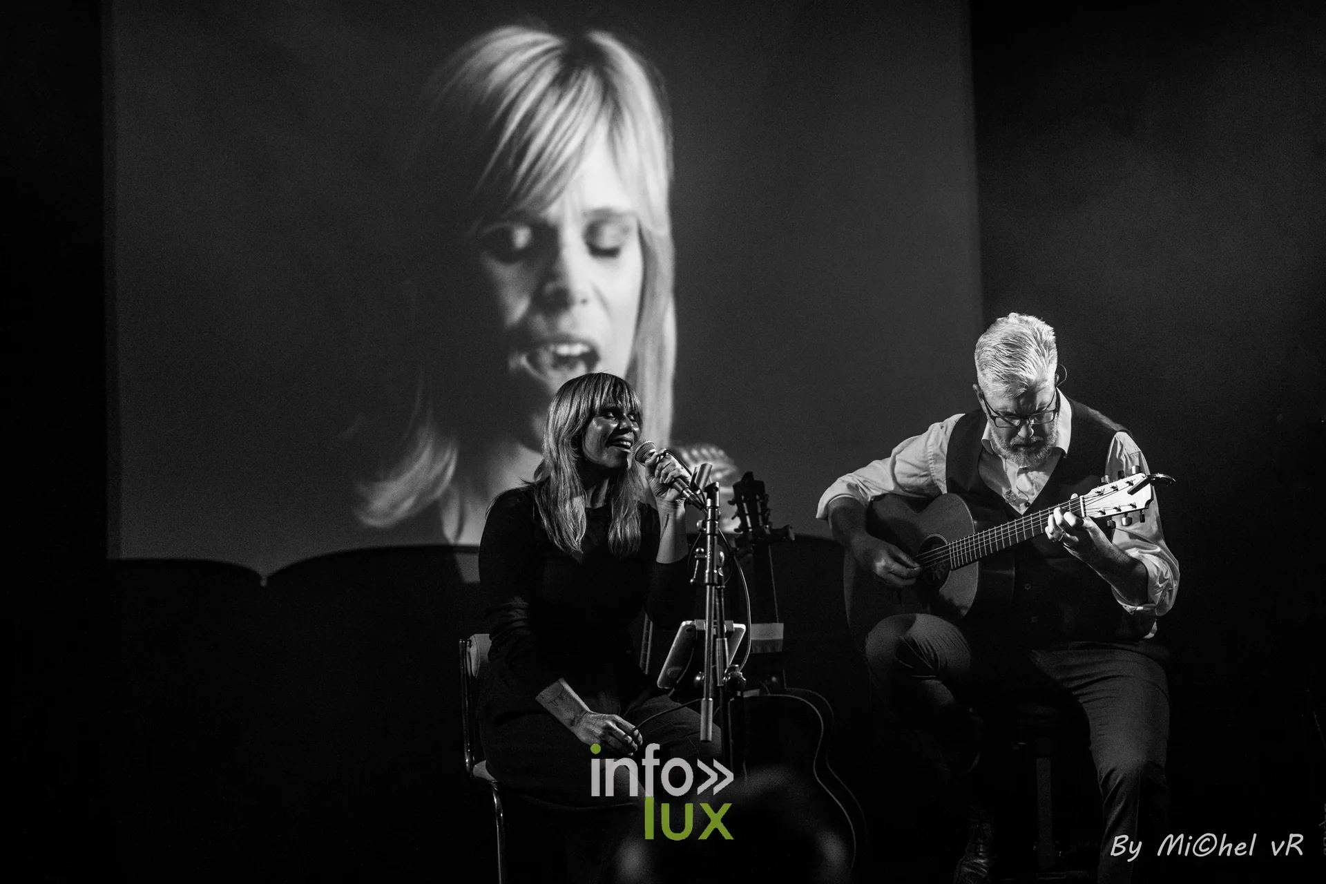 Le visage de Julie Compagnon projete en grand sur le fond de scene pendant que le duo joue devant, en noir et blanc