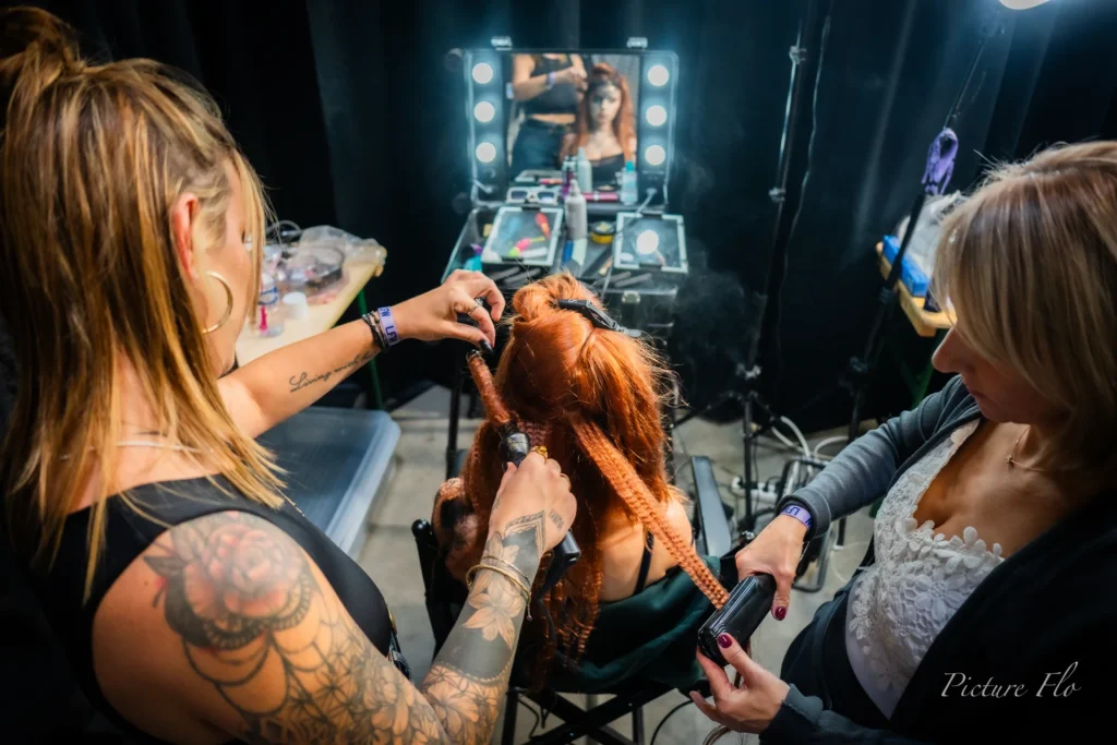 Deux coiffeuses travaillent une chevelure rousse devant un miroir lumineux, coulisses de la Lux Fashion Week 2026, Arlon