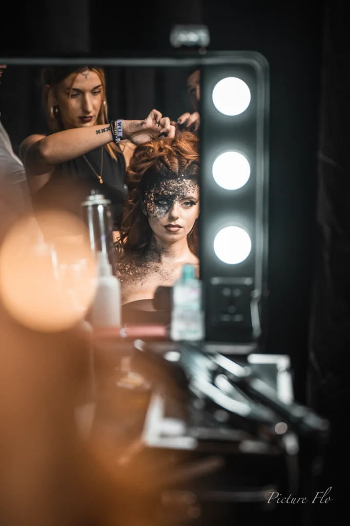Marine coiffée devant un miroir lumineux, maquillage noir et bronze, coulisses de la Lux Fashion Week 2026, Arlon