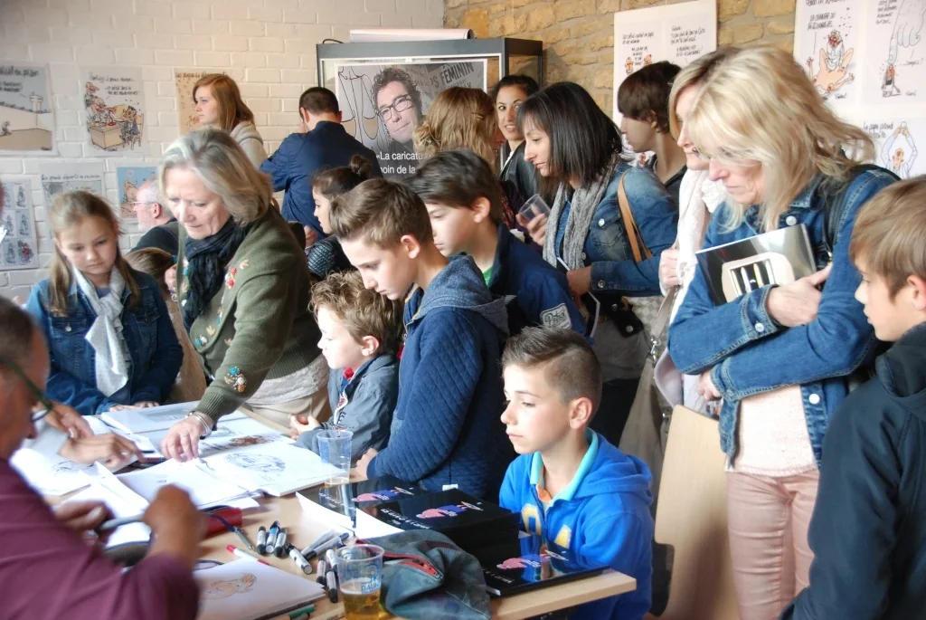 Groupe d’élèves et enseignante regardant un dessinateur travailler, dessins exposés au mur