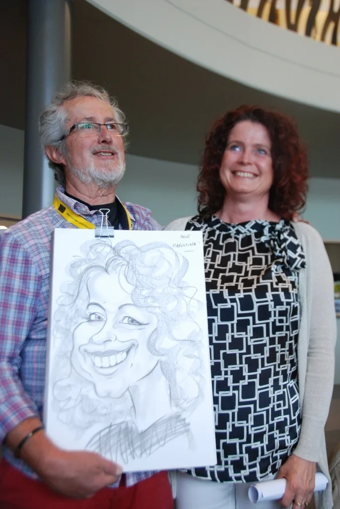 Un dessinateur et une visiteuse souriante posant avec la caricature qu’il vient de réaliser