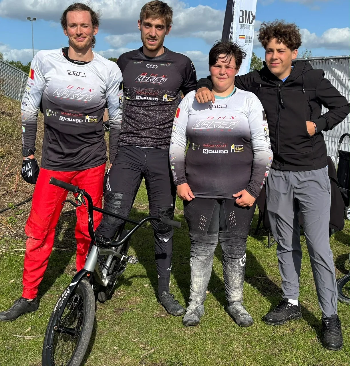 Quatre pilotes du BMX Timberwolves Racing Habay en maillot du club, un BMX au premier plan, devant la piste de Wachtebeke