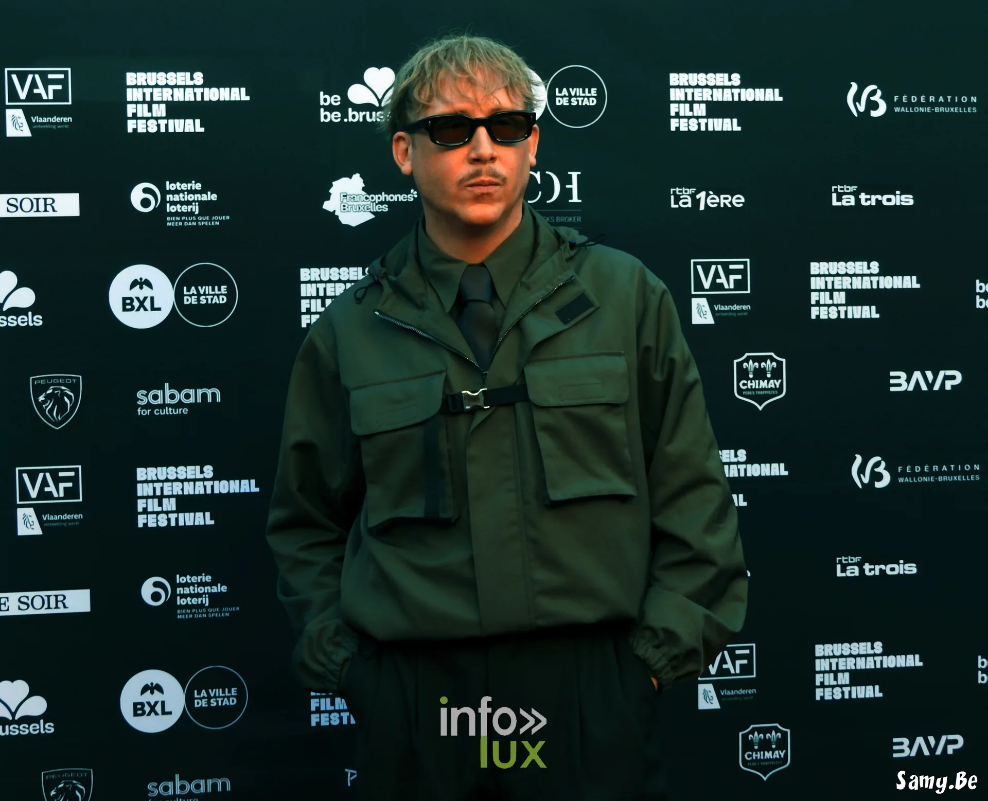 Un comédien de Vigilante en veste verte et lunettes noires au photocall