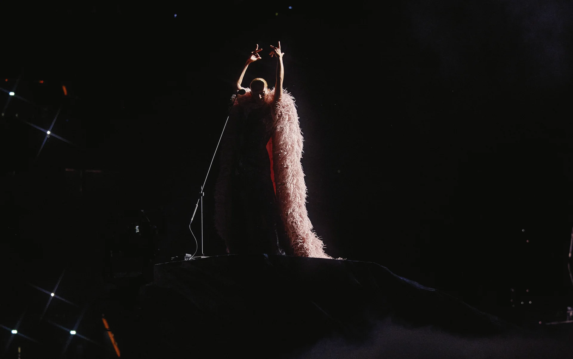Céline Dion en tenue Givenchy à plumes, bras levés, sur la scène sombre de la Plenitude Arena