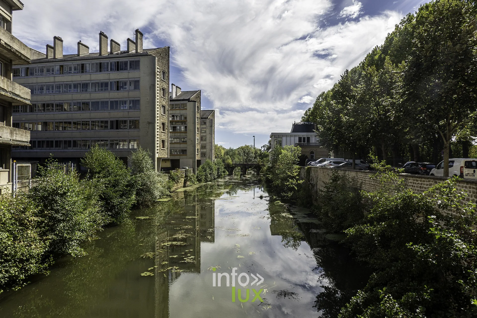 Bras d’eau bordé d’arbres et d’immeubles, reflets du ciel, Sedan