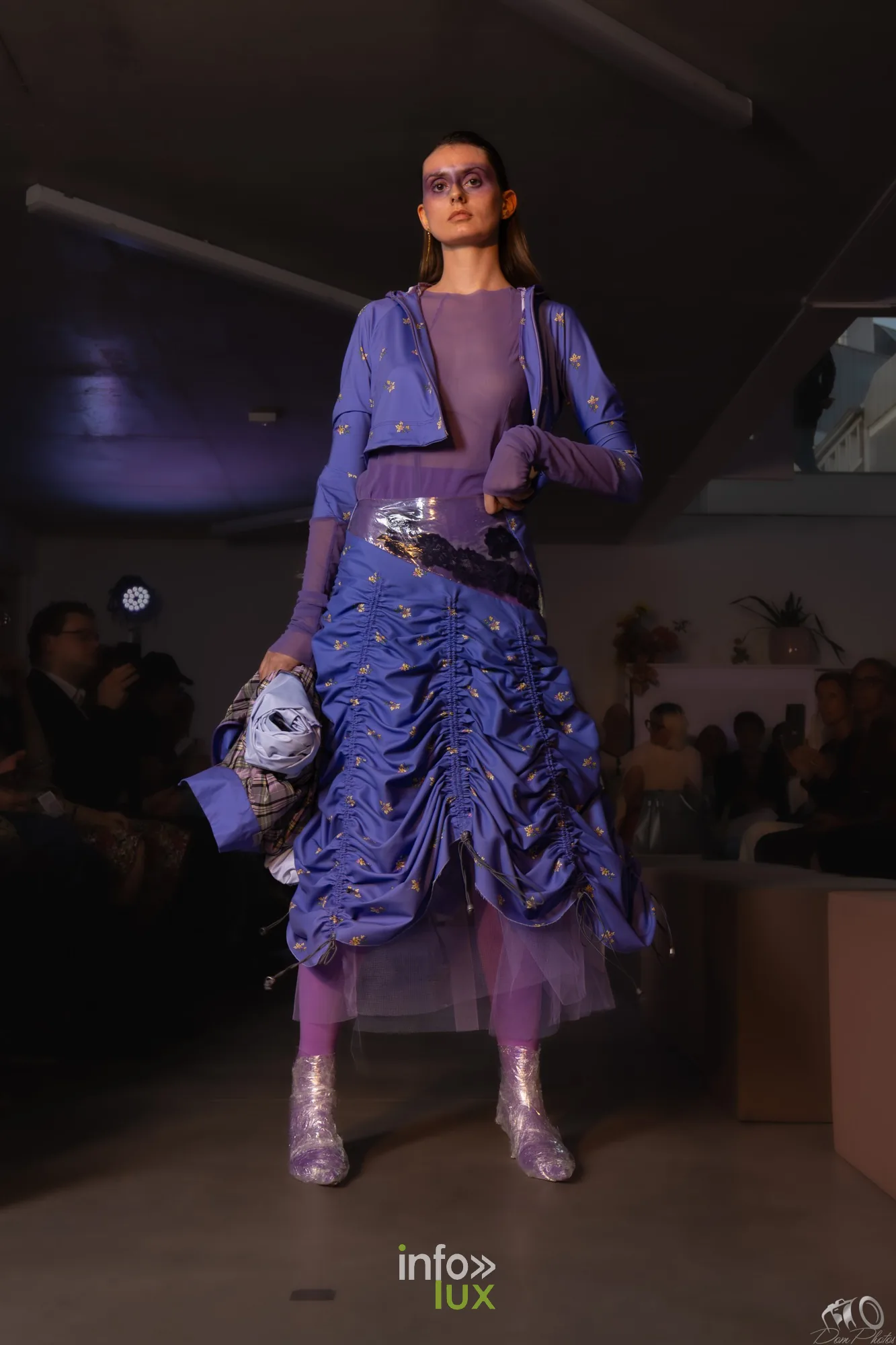 Mannequin en ensemble violet froncé, veste courte et jupe drapée, sac à main et bottes pailletées, devant le public