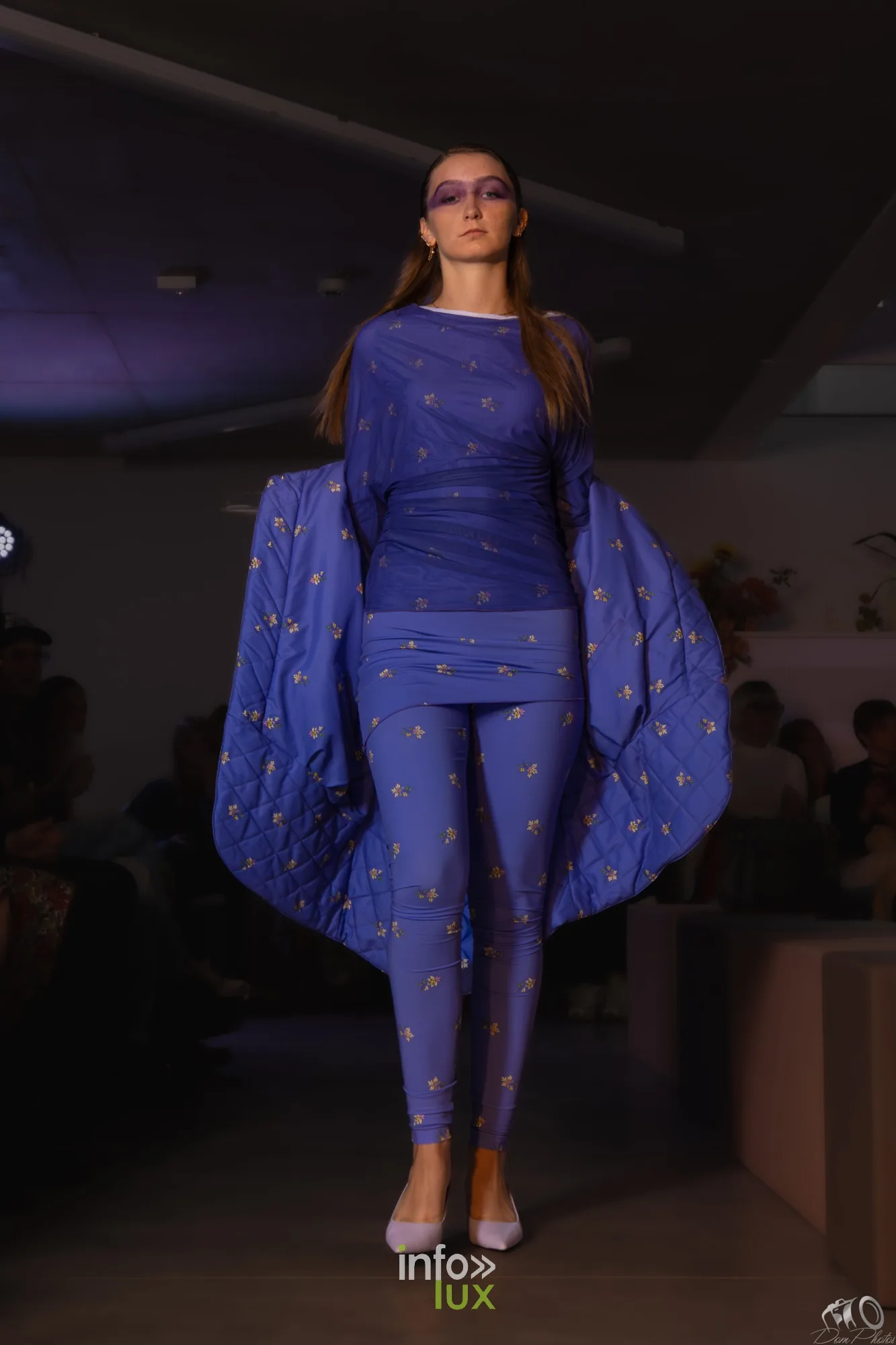 Mannequin en combinaison violette à motifs dorés avec une large manche-cape, sur le podium