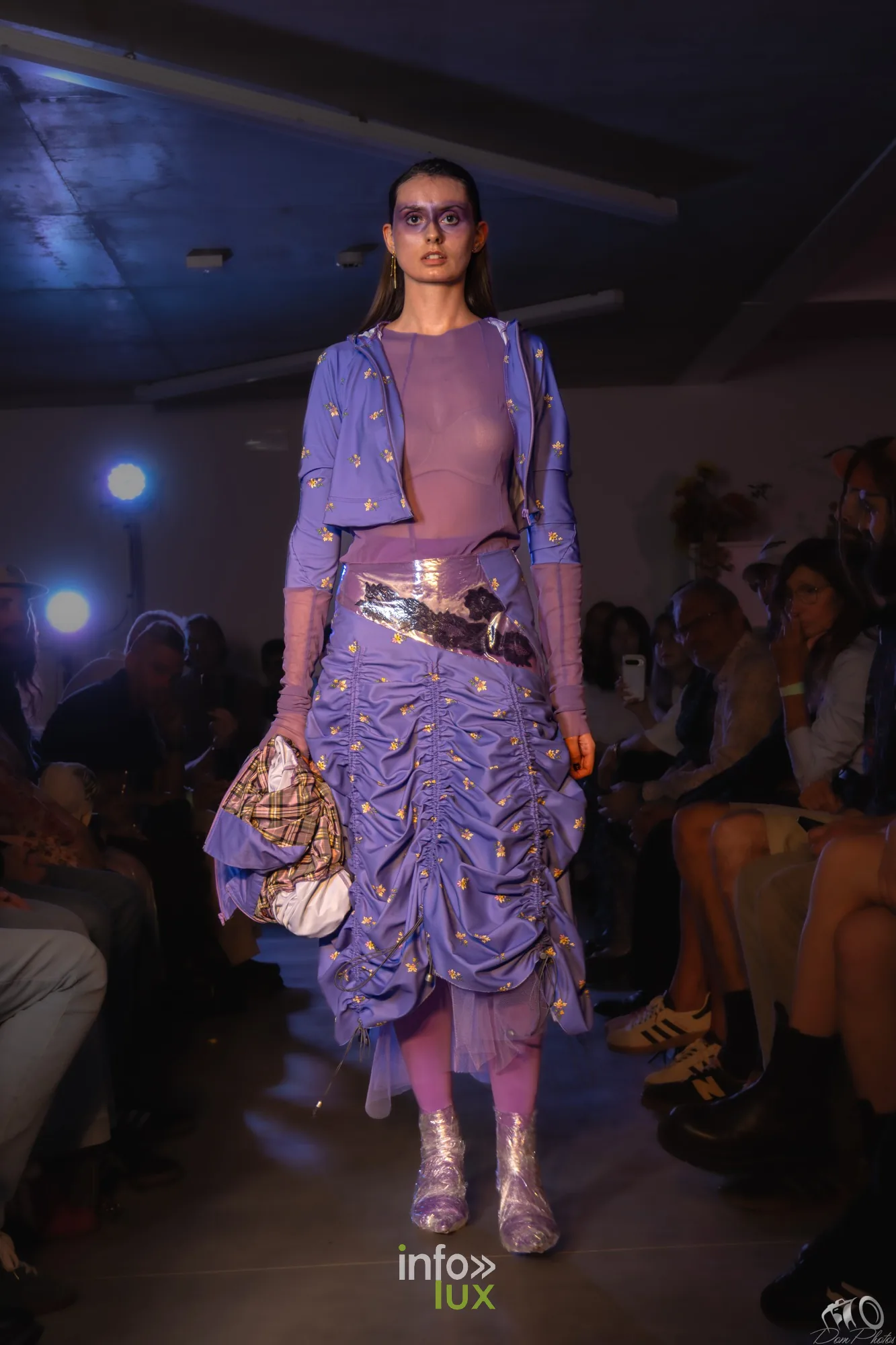 Mannequin en ensemble violet froncé, de face, avec un sac à main argenté, dans la lumière du podium