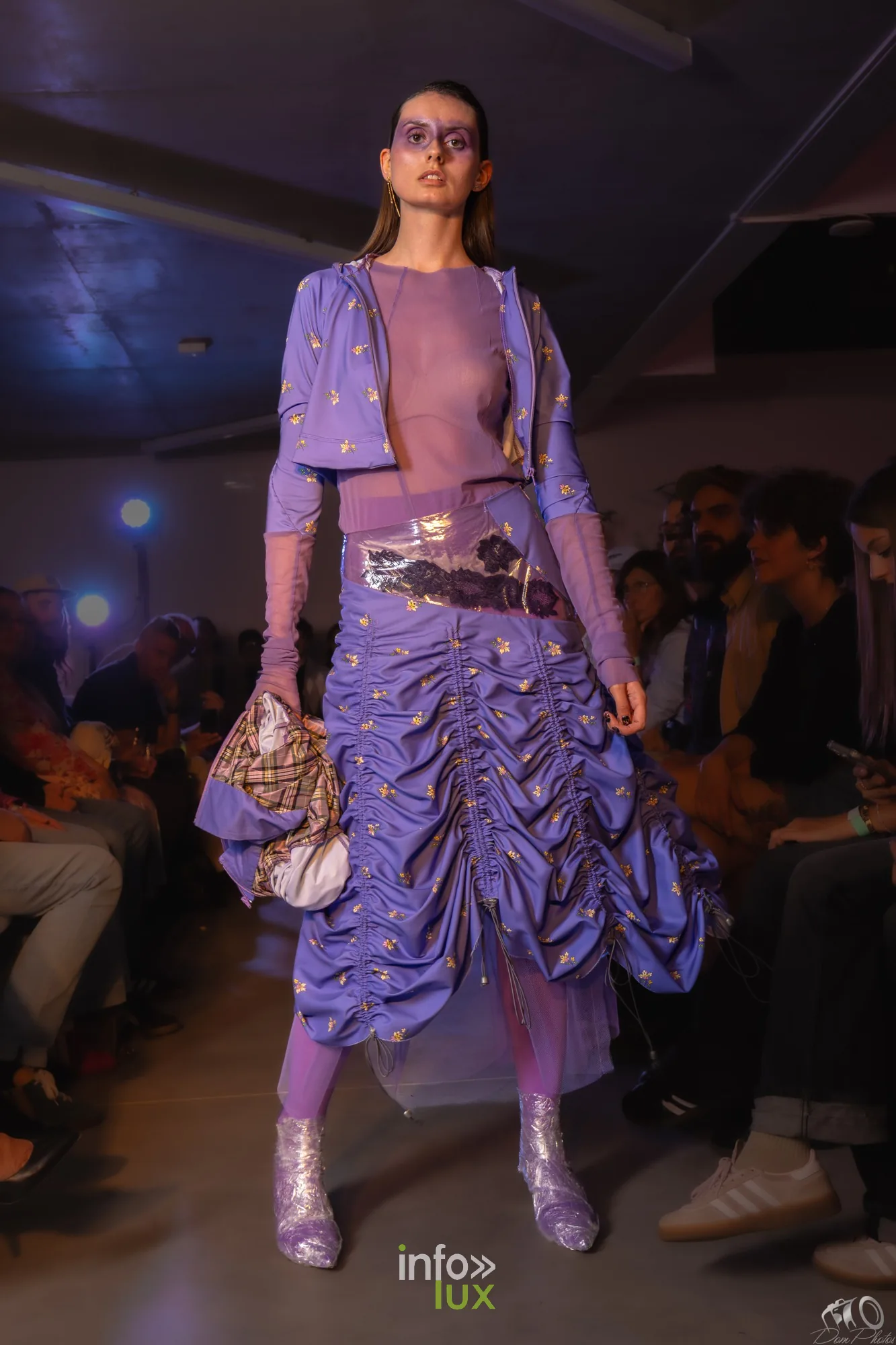 Mannequin en ensemble violet froncé, jupe drapée et bottes pailletées, devant les spectateurs assis