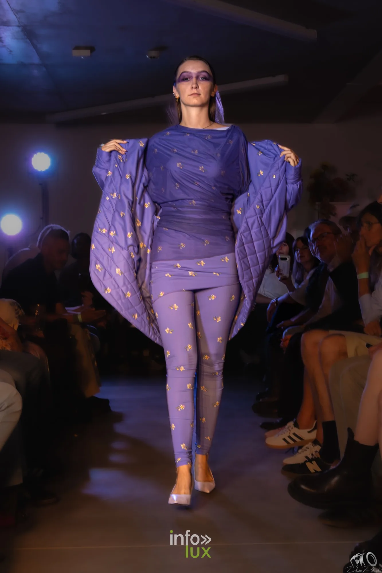 Mannequin en combinaison violette à motifs, bras écartés pour déployer une cape, sur le podium