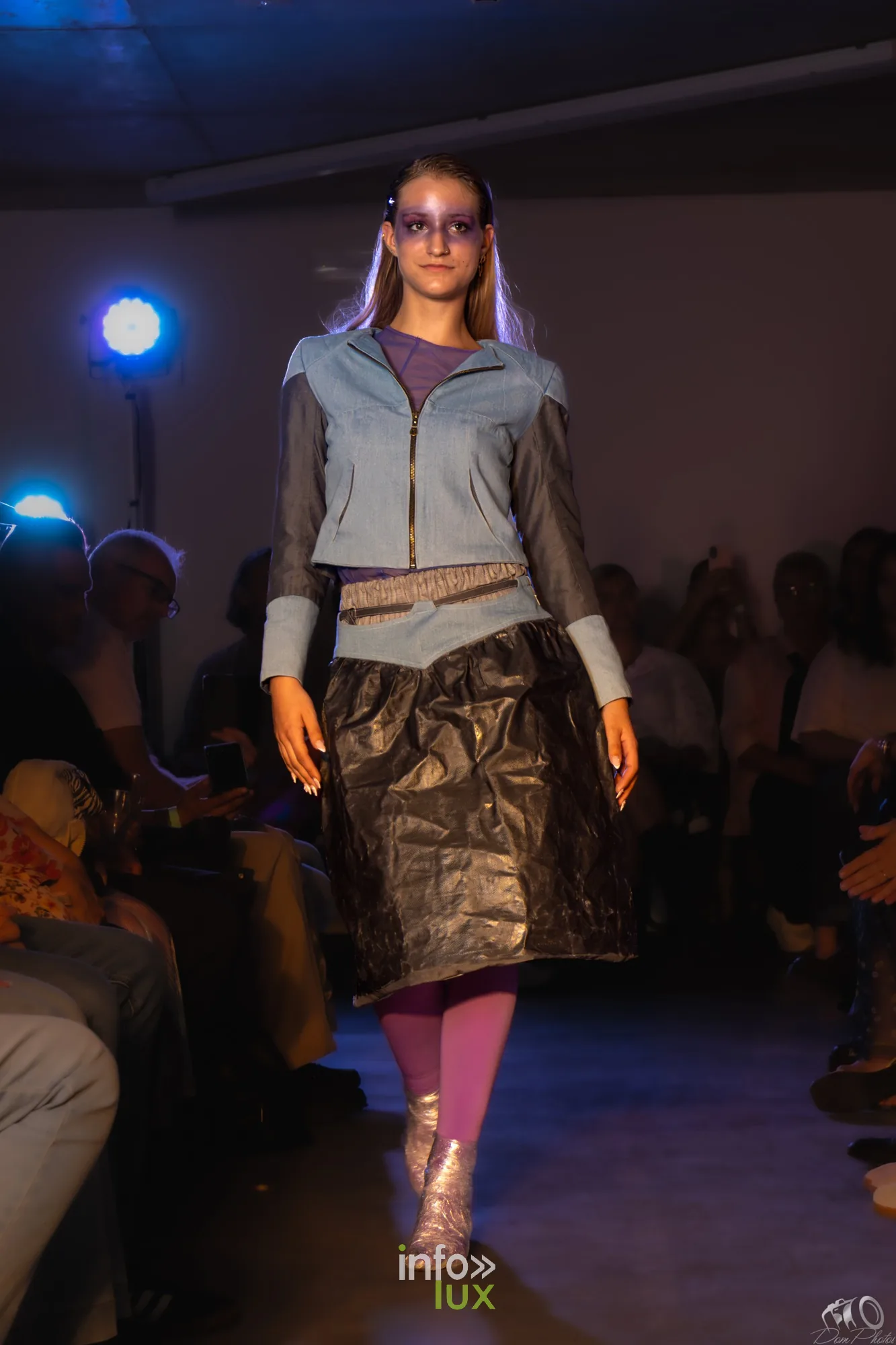 Mannequin en veste bleu ciel, jupe noire brillante et collants roses, sur le podium éclairé de bleu