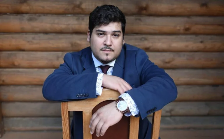 Le violoniste David Castro-Balbi en costume bleu, accoudé à une chaise devant un mur de bois