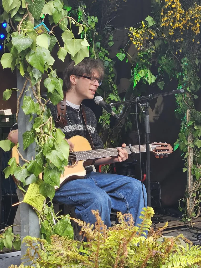 Jilo en concert, guitare acoustique à la main, devant un décor de feuillage