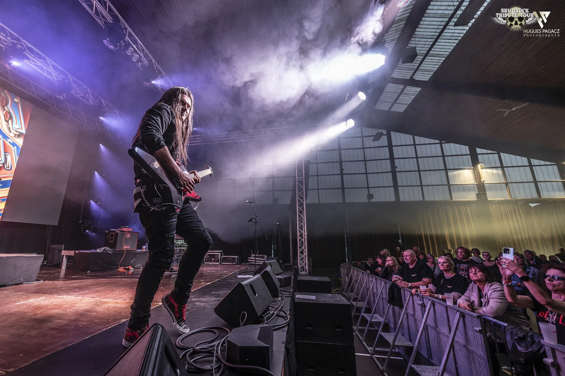 John L de dos face au public d’une salle de concert, dans la fumée et les projecteurs