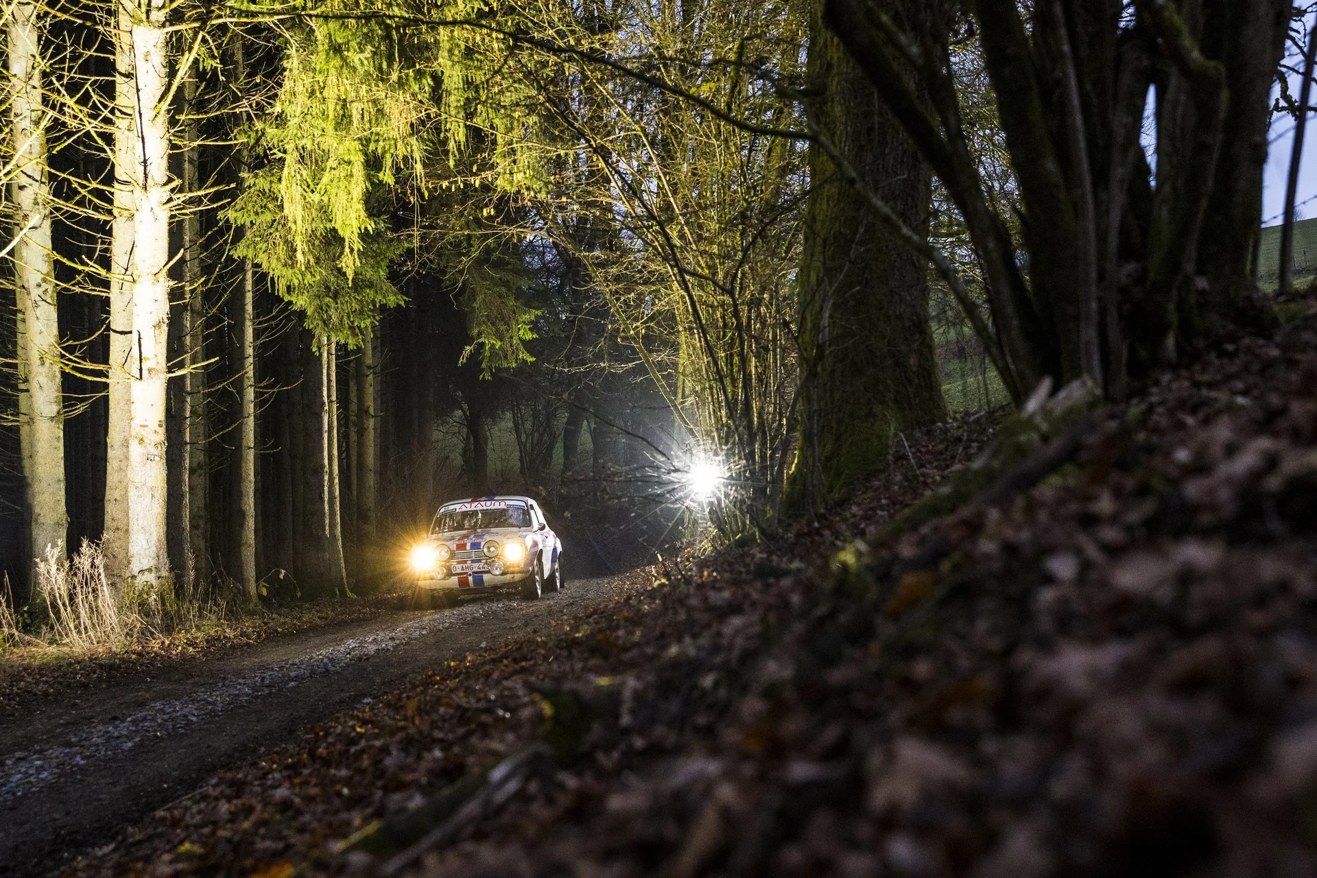 Une voiture de rallye historique phares allumés dans un chemin forestier, à la tombée du jour