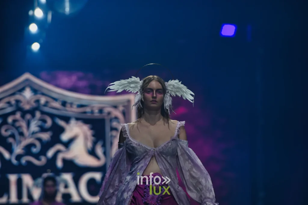 Chemise lilas ouverte à manches bouffantes, coiffe d’ailes blanches, devant le blason ALIMAC — Alimac, « Mystic Blossom », Lux Fashion Week 2026, Arlon