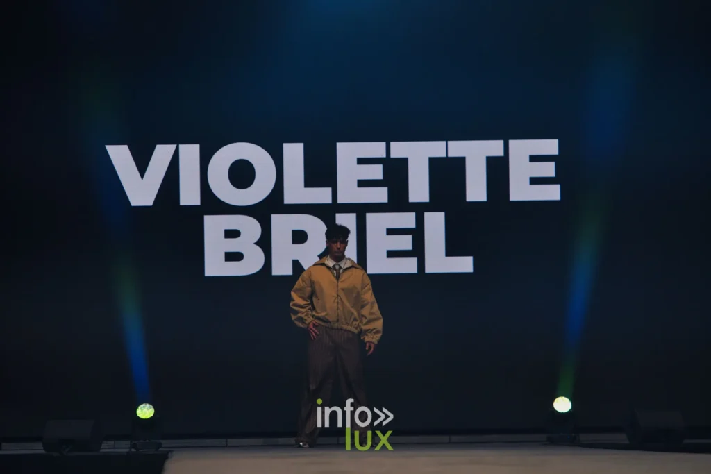 Le nom VIOLETTE BRIEL affiché sur l’écran de scène, un premier mannequin en blouson ocre devant — Violette Briel, « L’Héritage », Lux Fashion Week 2026, Arlon