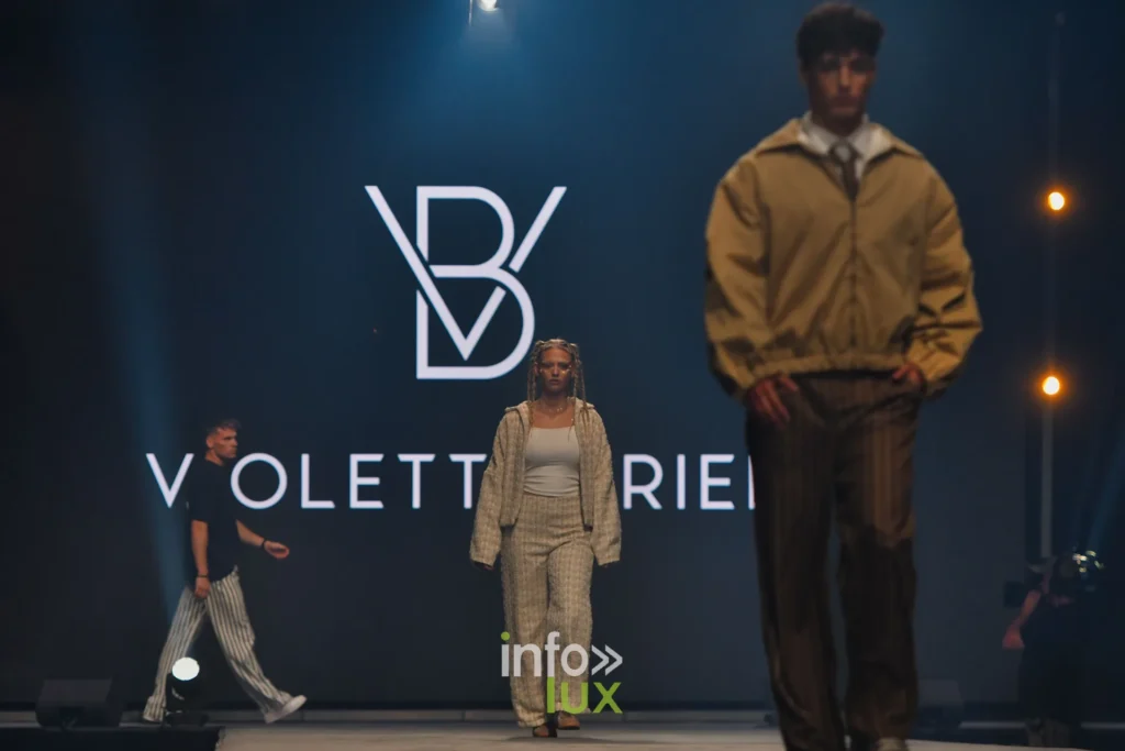 Blouson ocre et pantalon brun à fines rayures, devant le monogramme VB de la créatrice à l’écran — Violette Briel, « L’Héritage », Lux Fashion Week 2026, Arlon