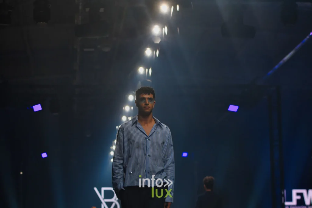 Chemise bleue à rayures et pantalon noir, le monogramme VB à l’écran — Violette Briel, « L’Héritage », Lux Fashion Week 2026, Arlon