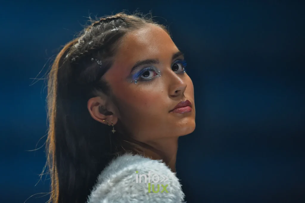 Portrait : cheveux tressés, maquillage bleu pailleté, col blanc à longs poils — Violette Briel, « L’Héritage », Lux Fashion Week 2026, Arlon