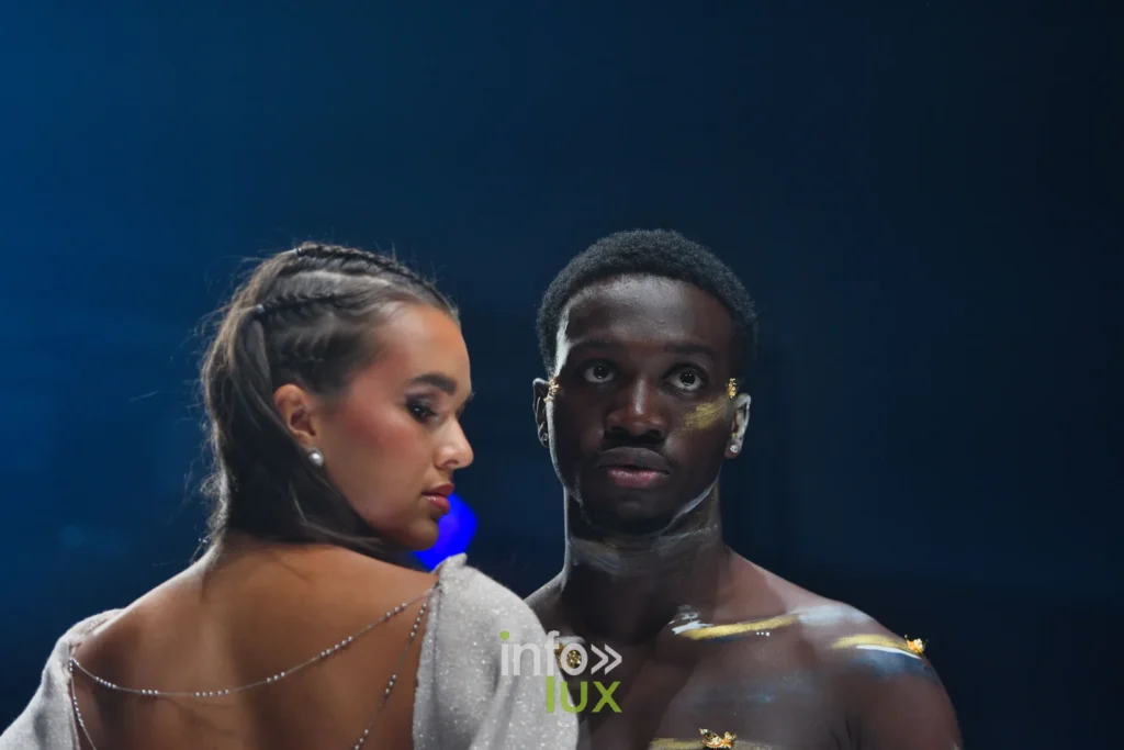 Robe argentée scintillante au dos nu traversé de chaînettes ; à côté, un mannequin torse nu au maquillage doré — Violette Briel, « L’Héritage », Lux Fashion Week 2026, Arlon