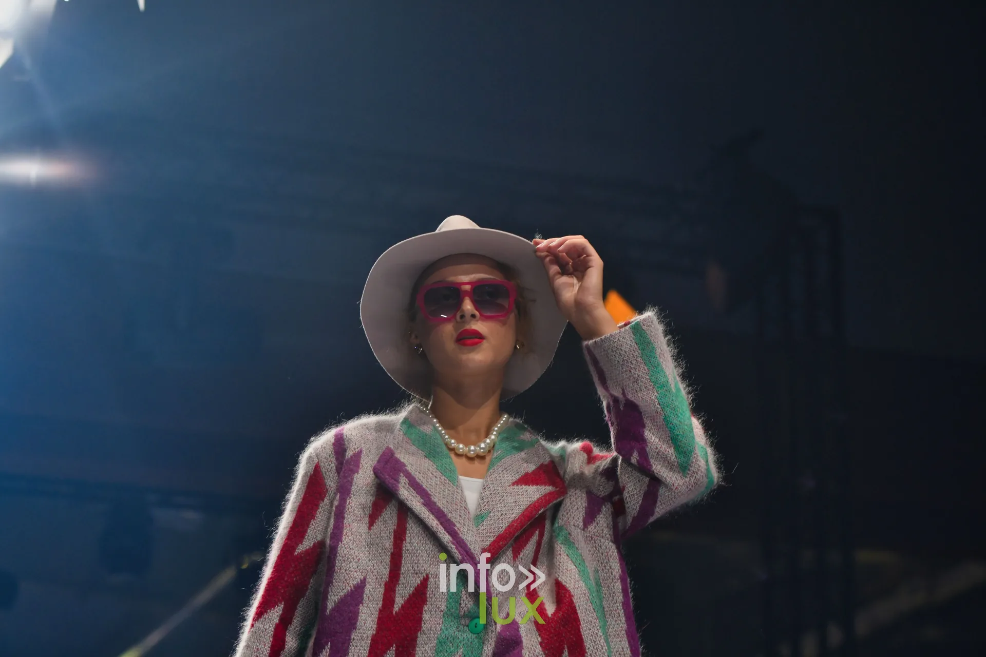 Manteau en maille gris à éclairs rouges, violets et verts, main posée sur le chapeau blanc — Damlaws, « The Dam Star », Lux Fashion Week 2026, Arlon