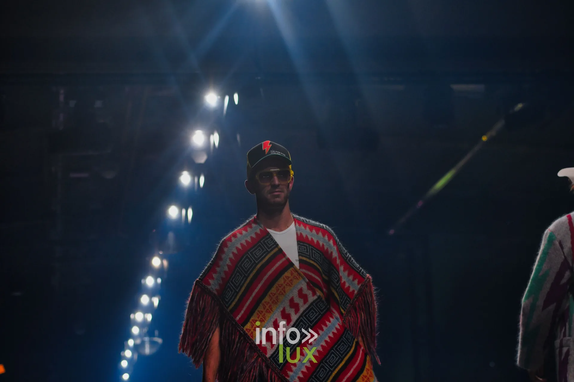 Poncho à franges rouge, noir et jaune à motifs géométriques, casquette noire à éclair rouge — Damlaws, « The Dam Star », Lux Fashion Week 2026, Arlon