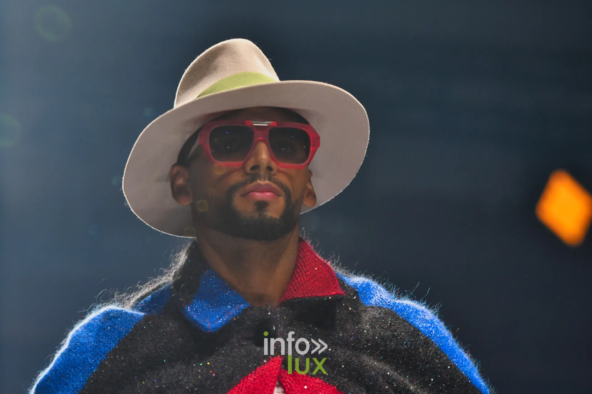 Cape en maille noire à chevrons rouge et bleu, col rouge, chapeau blanc et lunettes rouges — Damlaws, « The Dam Star », Lux Fashion Week 2026, Arlon