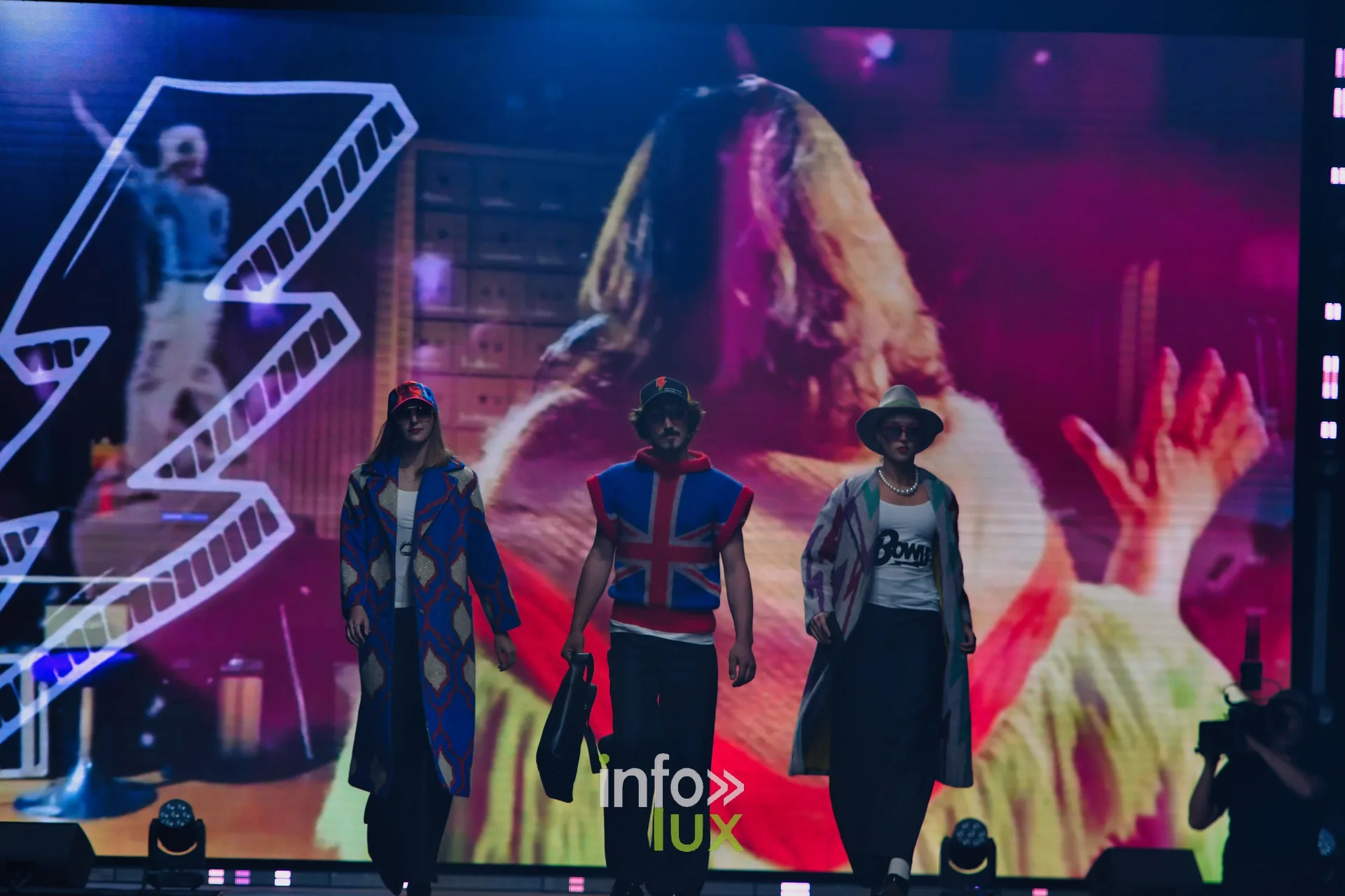 Trois silhouettes devant l’écran où s’affiche un éclair dessiné ; t-shirt imprimé « Bowie » à droite, pull Union Jack au centre — Damlaws, « The Dam Star », Lux Fashion Week 2026, Arlon