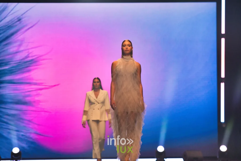 Robe longue en tulle à volants disposés en losanges, col montant, devant un écran aux dégradés rose et bleu ; derrière, un tailleur-pantalon crème — Francini_K, « La géométrie des fleurs », Lux Fashion Week 2026, Arlon