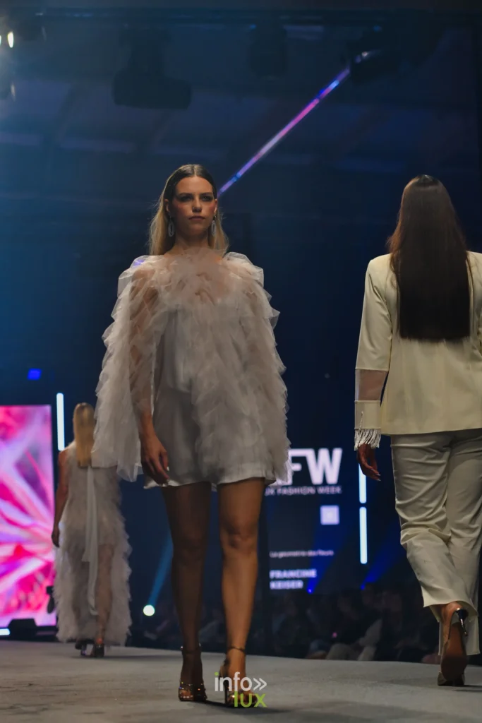 Robe courte en satin blanc sous une cape de tulle à volants ; à l’écran, « La géométrie des fleurs » et le nom de Francine Keiser — Francini_K, « La géométrie des fleurs », Lux Fashion Week 2026, Arlon