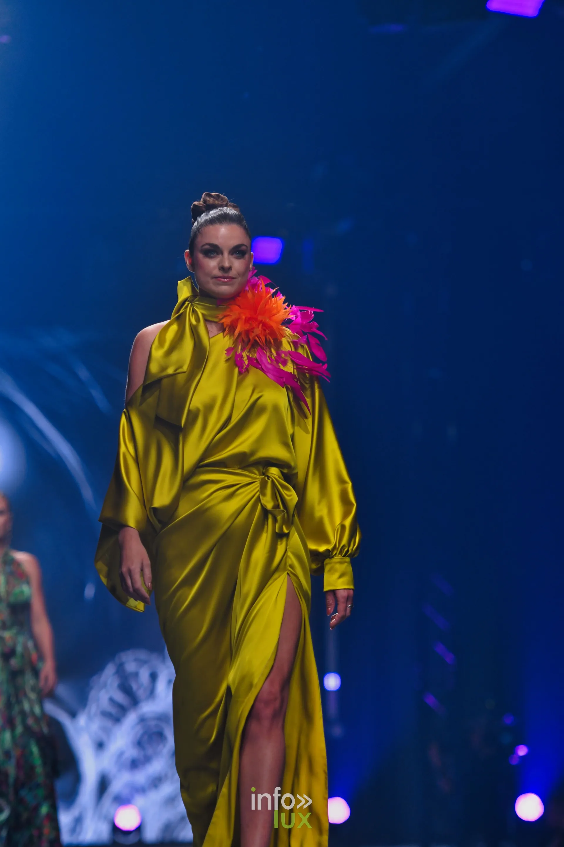 Robe drapée en satin jaune, fendue, plumes orange et fuchsia à l’épaule, collection « Meraki bloom » d’Hélène Guiot, Lux Fashion Week 2026, Arlon