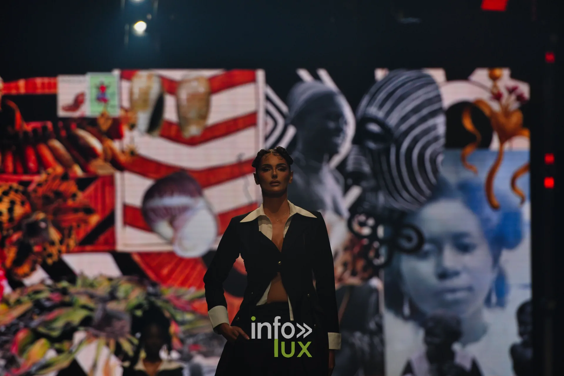 Tailleur noir à col et poignets blancs devant l’écran : collage de motifs graphiques rouge et blanc et de portraits en noir et blanc — R. Lopes, « Raízes d’Ouro », Lux Fashion Week 2026, Arlon