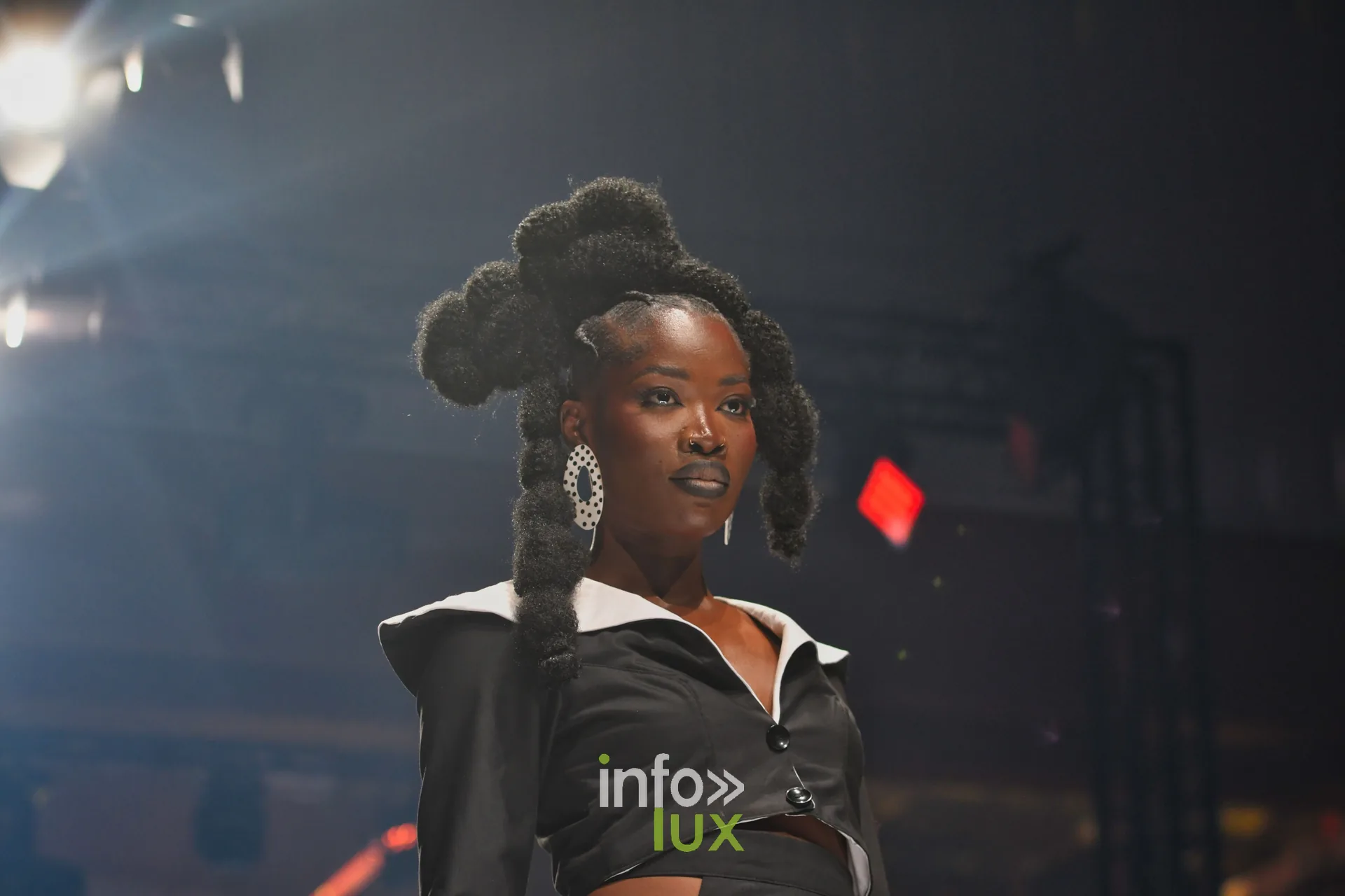 Veste courte noire à col blanc, coiffure en chignons sculptés, grandes boucles d’oreilles — R. Lopes, « Raízes d’Ouro », Lux Fashion Week 2026, Arlon