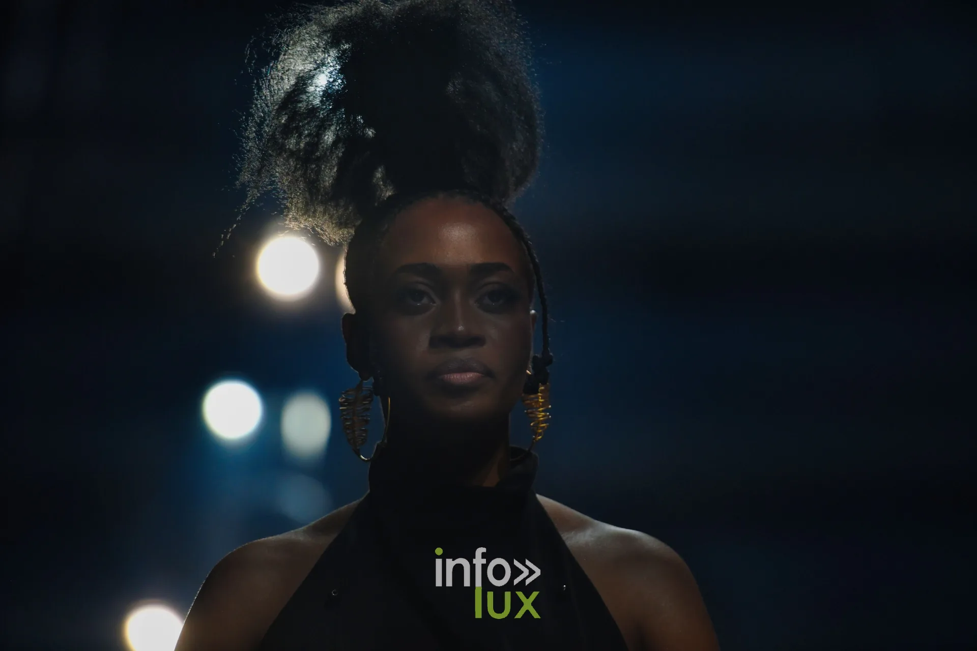 Haut noir à col montant, chignon volumineux et longues boucles dorées, à contre-jour — R. Lopes, « Raízes d’Ouro », Lux Fashion Week 2026, Arlon