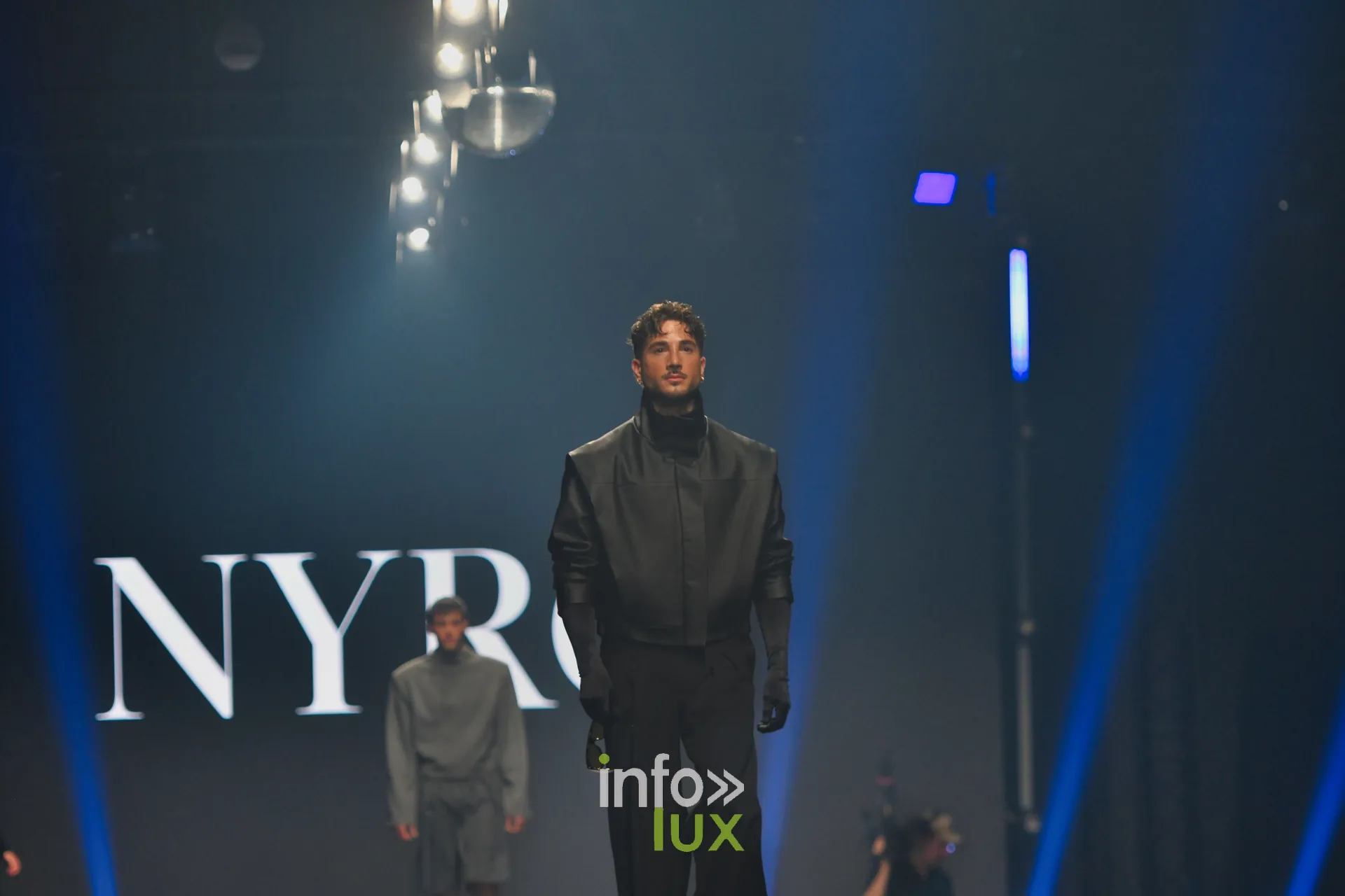 Veste sans manches en cuir noir à col montant et gants noirs, devant l’écran « NYRO », collection « NYRO » de Mathieu Panzokou, Lux Fashion Week 2026, Arlon