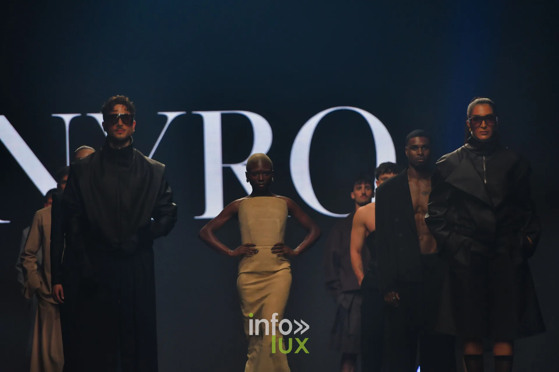 Silhouettes alignées devant l’écran « NYRO » : robe bustier longue beige, manteaux noirs, lunettes noires, collection « NYRO » de Mathieu Panzokou, Lux Fashion Week 2026, Arlon
