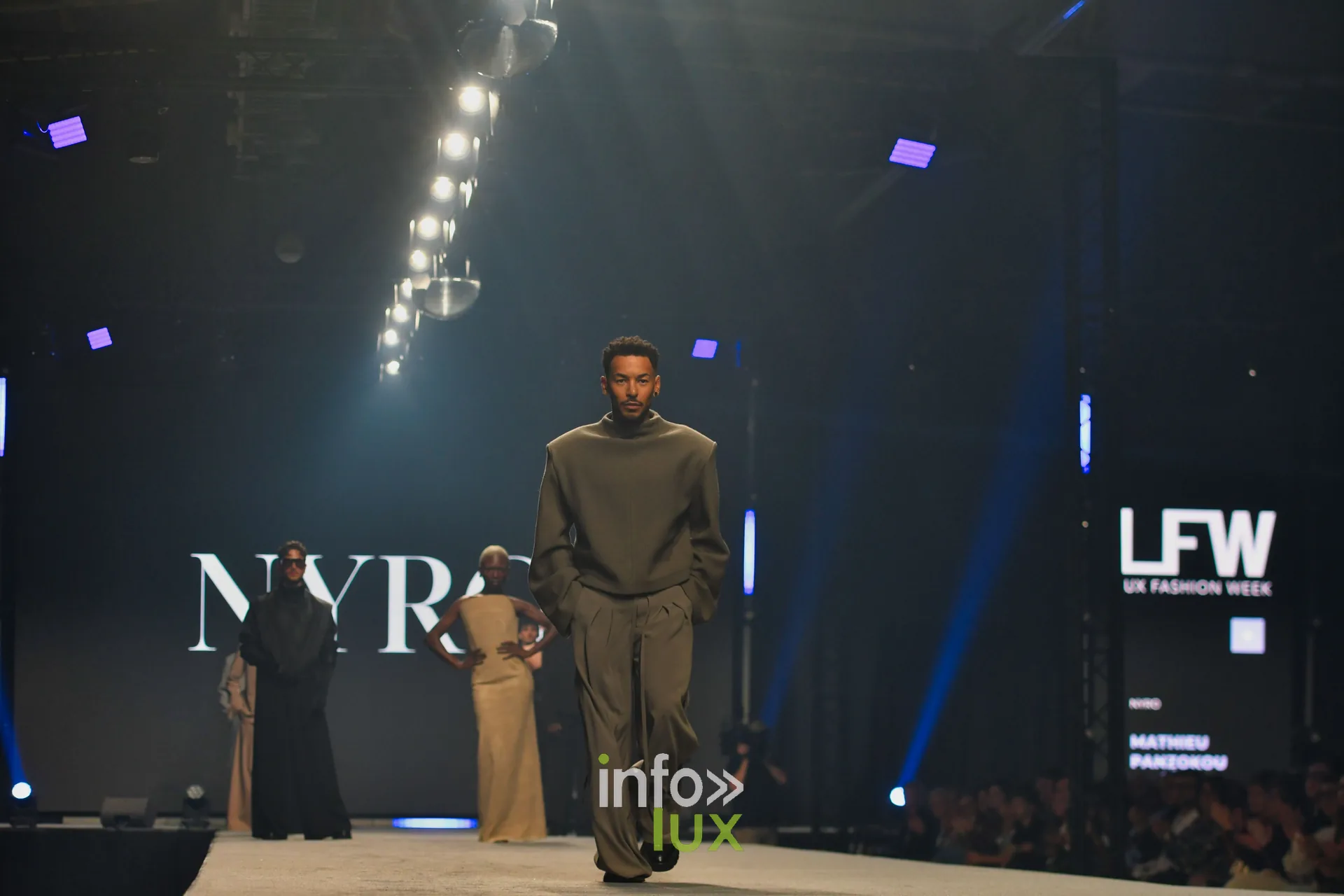 Vue du podium : ensemble gris-vert à épaules structurées, écran « NYRO » en fond, collection « NYRO » de Mathieu Panzokou, Lux Fashion Week 2026, Arlon