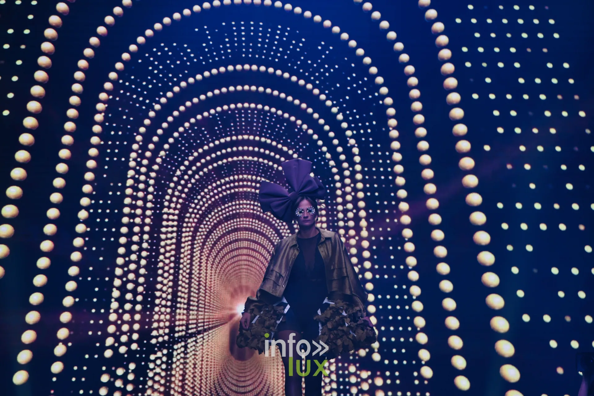 Première silhouette sous l’arche lumineuse projetée sur l’écran : nœud violet géant, veste dorée à volants — Charlotte Spoidenne, « Diffraction », Lux Fashion Week 2026, Arlon