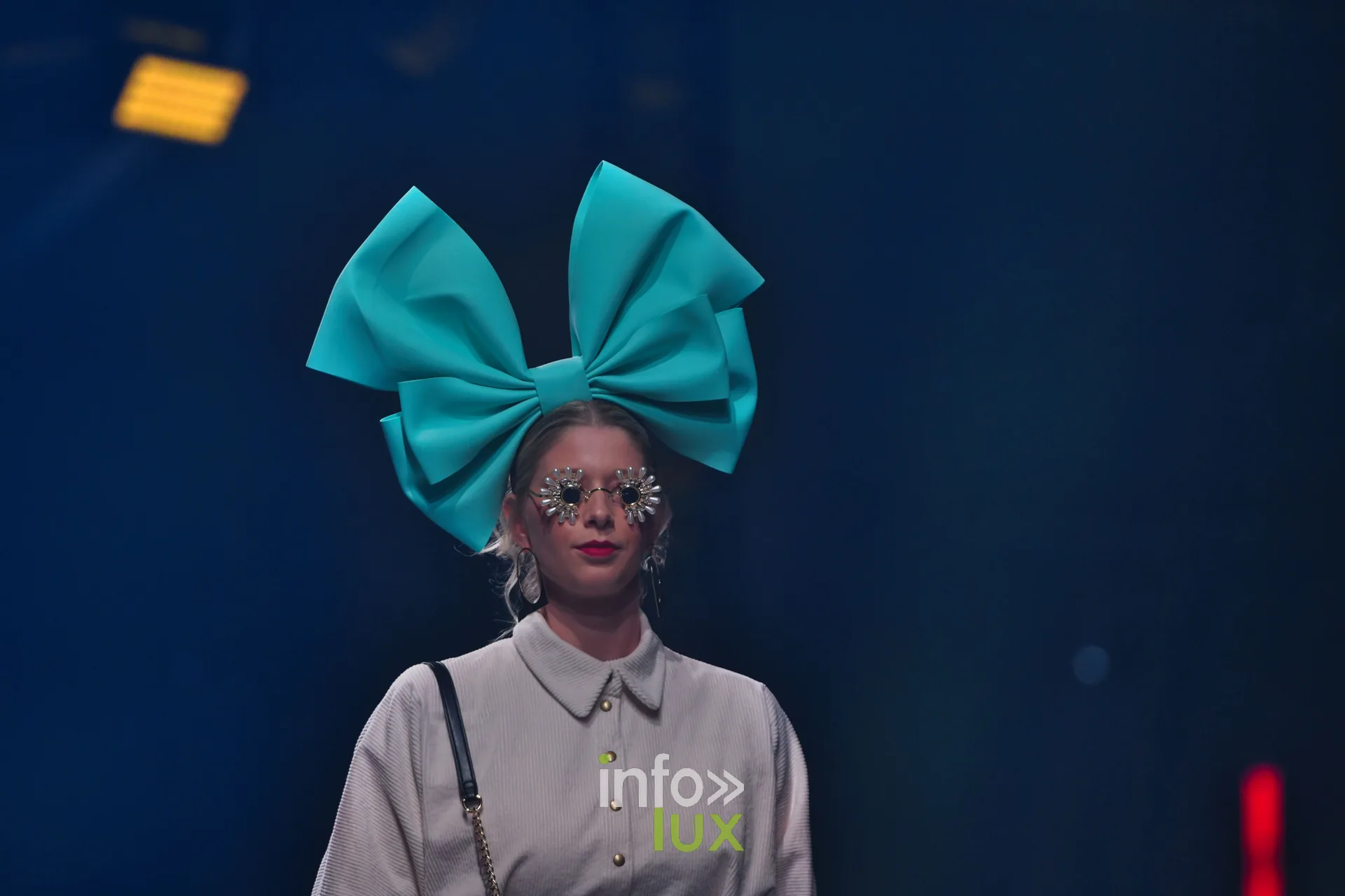 Nœud turquoise géant, chemise grise à boutons dorés — Charlotte Spoidenne, « Diffraction », Lux Fashion Week 2026, Arlon