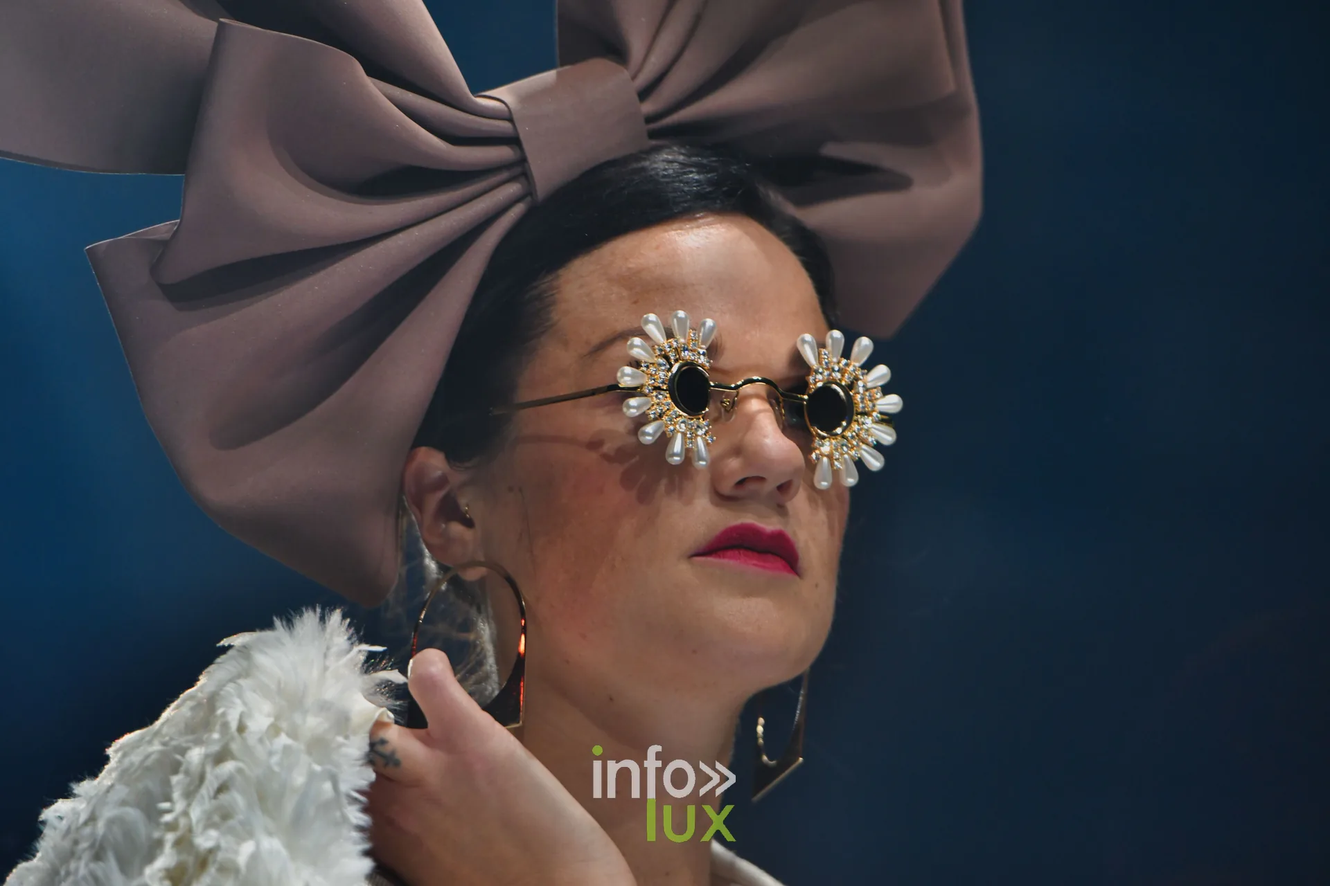 Nœud géant taupe, lunettes ornées de fleurs et plumes blanches, en gros plan — Charlotte Spoidenne, « Diffraction », Lux Fashion Week 2026, Arlon