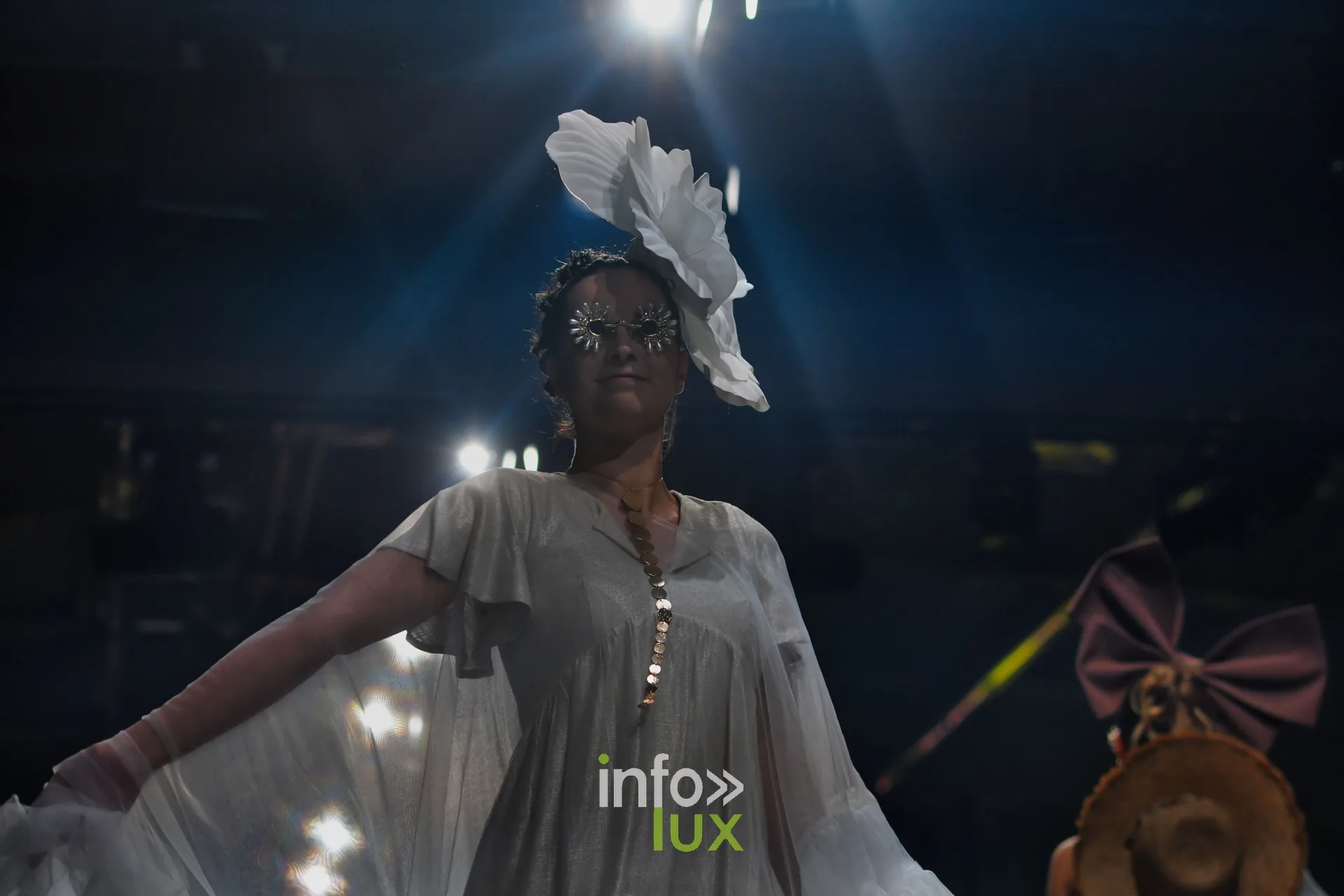 Robe grise fluide à boutons dorés, bras écartés, grande fleur blanche en coiffe — Charlotte Spoidenne, « Diffraction », Lux Fashion Week 2026, Arlon