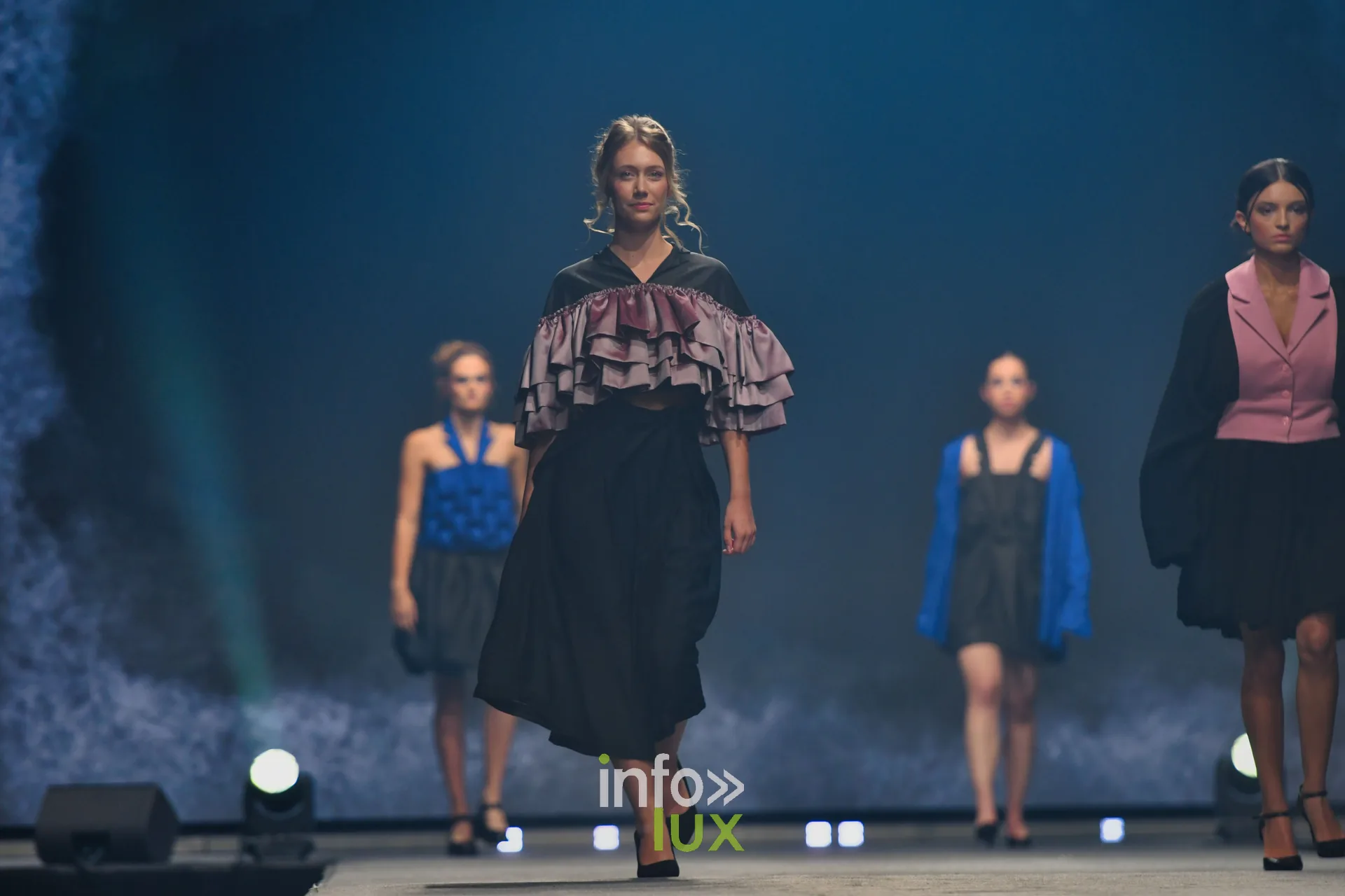Passage de groupe : cape à volants rose sur jupe noire au premier plan, haut bleu roi, gilet bleu et gilet rose derrière — Victoire Waxweiler, « Fragments », Lux Fashion Week 2026, Arlon