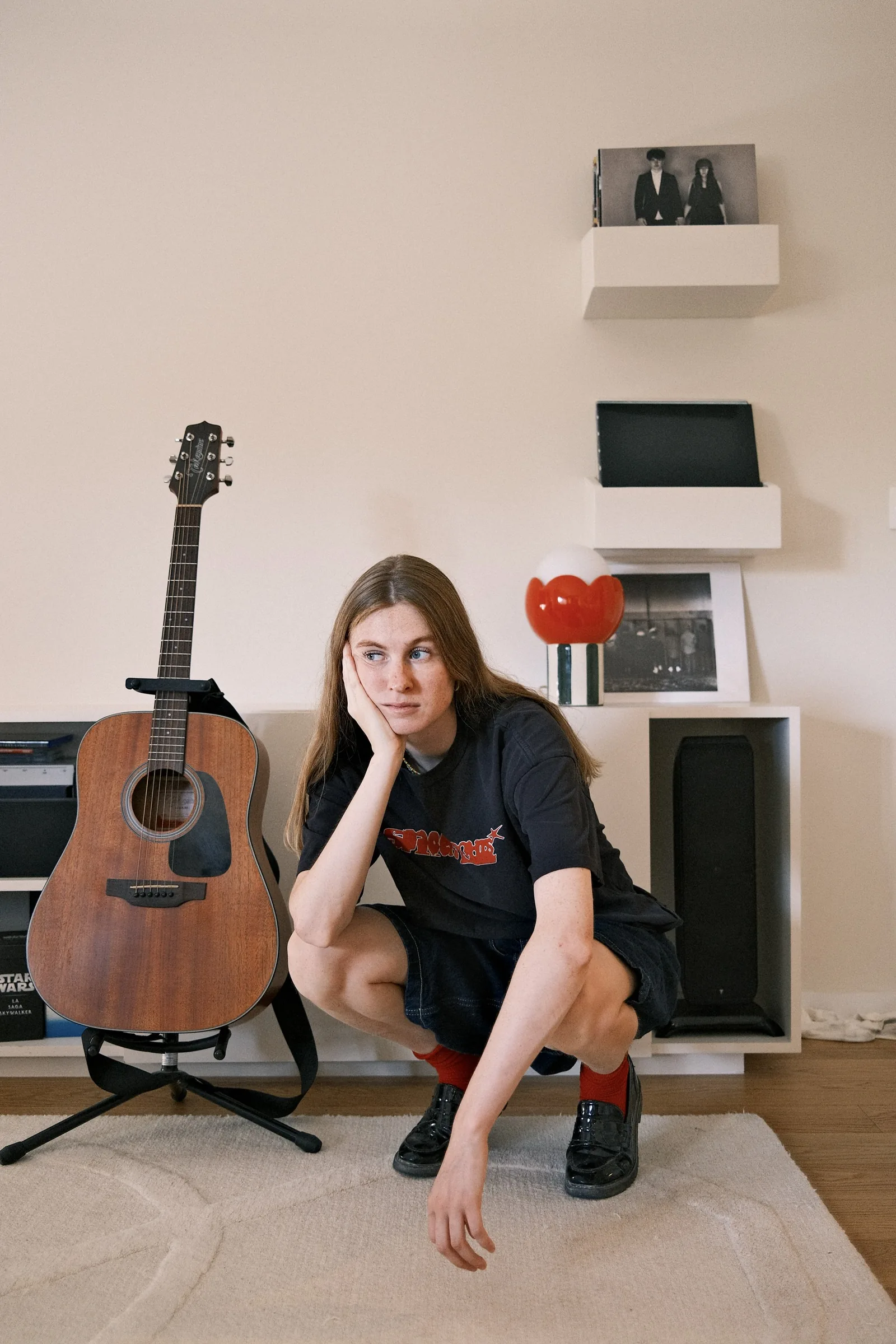 Maguie accroupie dans une pièce claire, une guitare acoustique à côté d’elle