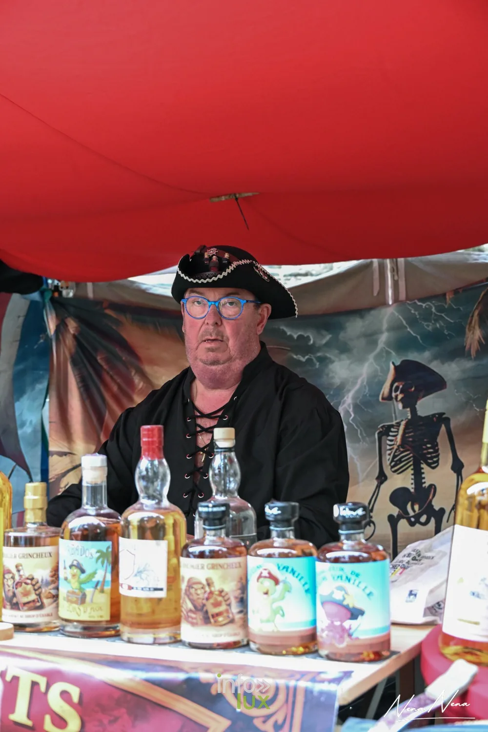 Un marchand en tricorne derrière ses bouteilles d’élixirs, sous une tente rouge du marché médiéval