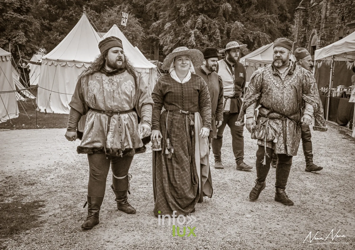 Un groupe de médiévistes en costume marche entre les tentes du campement, en sépia