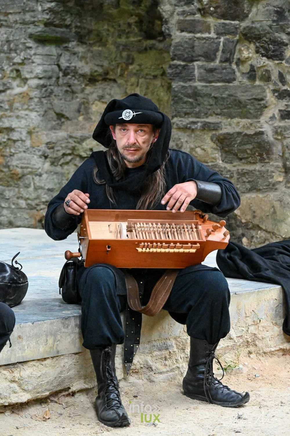 Un musicien en noir joue de la vielle à roue, assis sur un muret de pierre